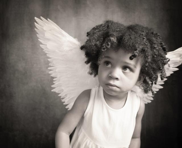 Black and white photograph. A young girl with angelic wings gazes towards the left.