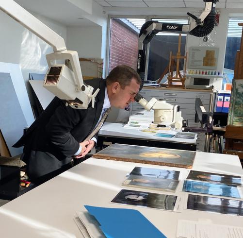 Photograph of a man in a suit looking through a microscope at a painting