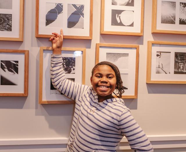 Photograph of a student standing in front of a wall of framed prints, pointing at a print